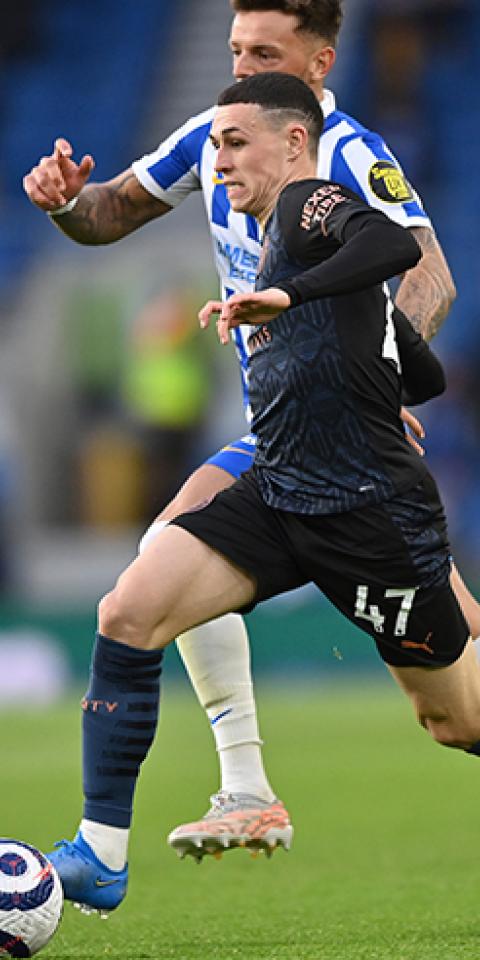 Phil Foden controla un balón perseguido por un rival. Conoce las cuotas y pronósticos del Manchester City Vs Everton.